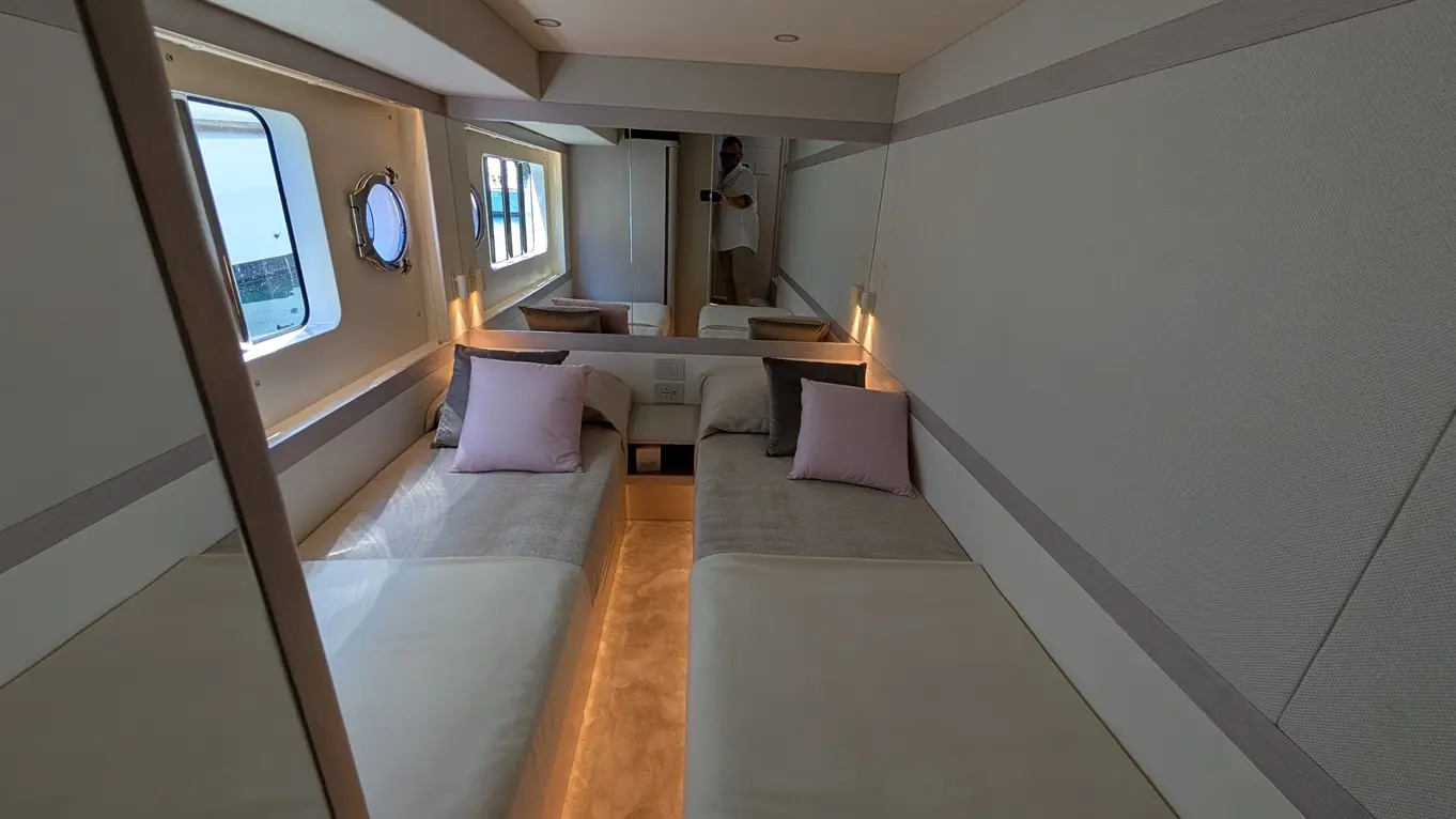 Brightly lit cabin with two single beds, decorative pillows, and a mirror, featuring a window and neutral decor.