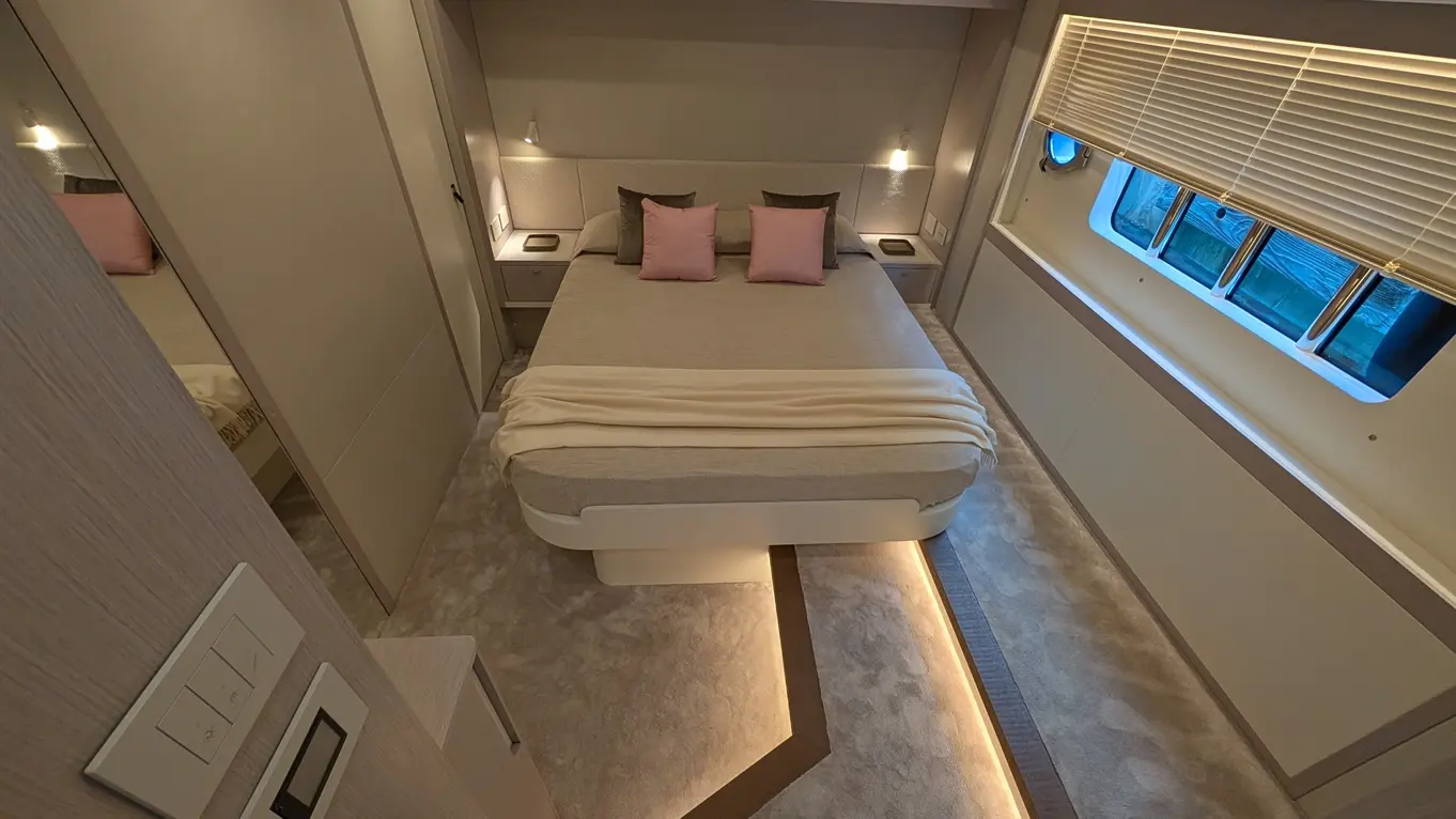 Modern bedroom with a double bed, pink cushions, warm lighting, and a large window featuring blinds by a carpeted floor.