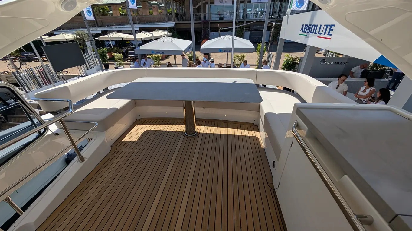 Spacious yacht deck with wooden flooring, a grey table, and white seating, surrounded by sun umbrellas and attendees.
