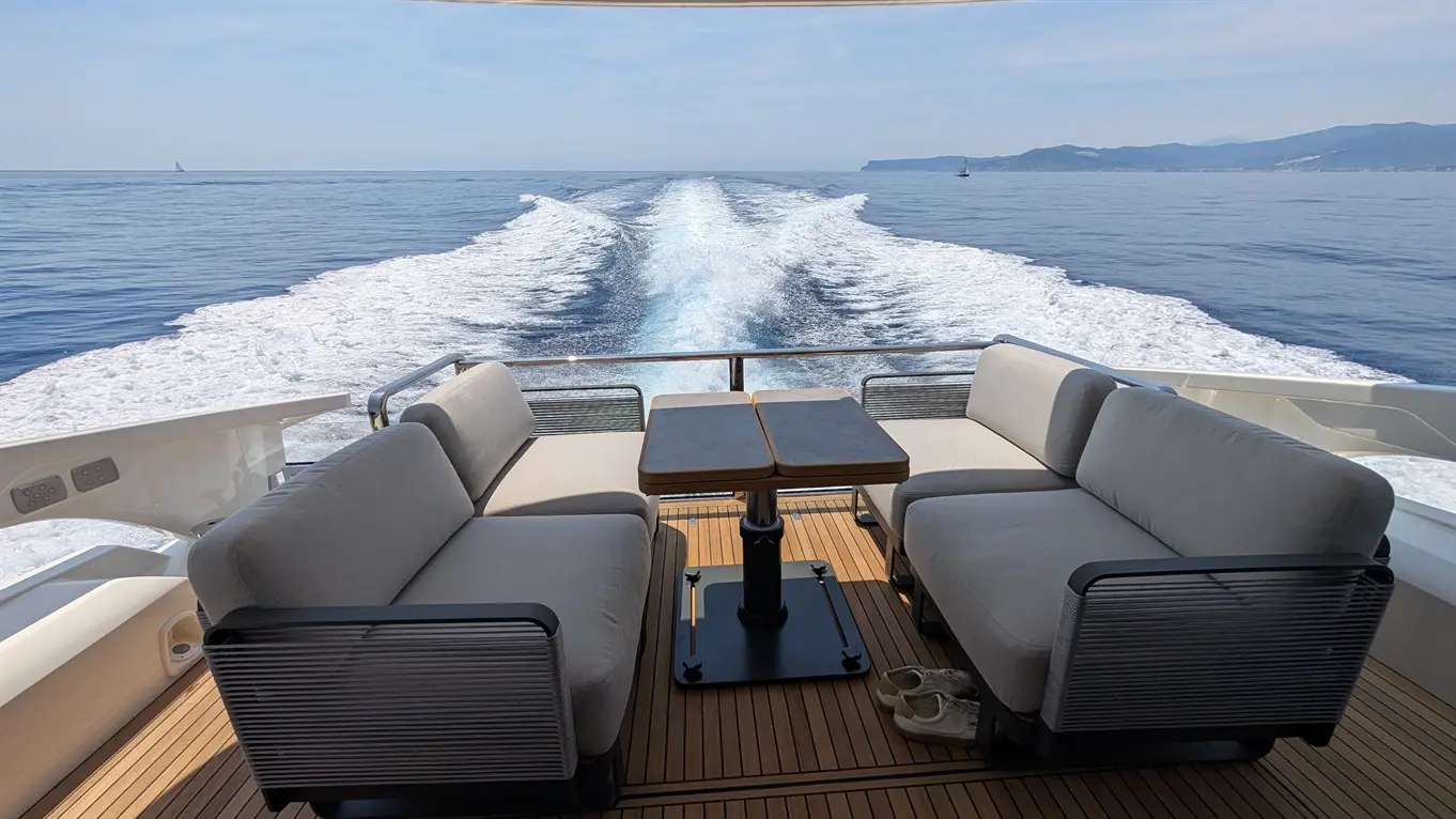 Luxury yacht deck with light grey sofas and a coffee table, overlooking the tranquil sea and wake against a distant coastline.