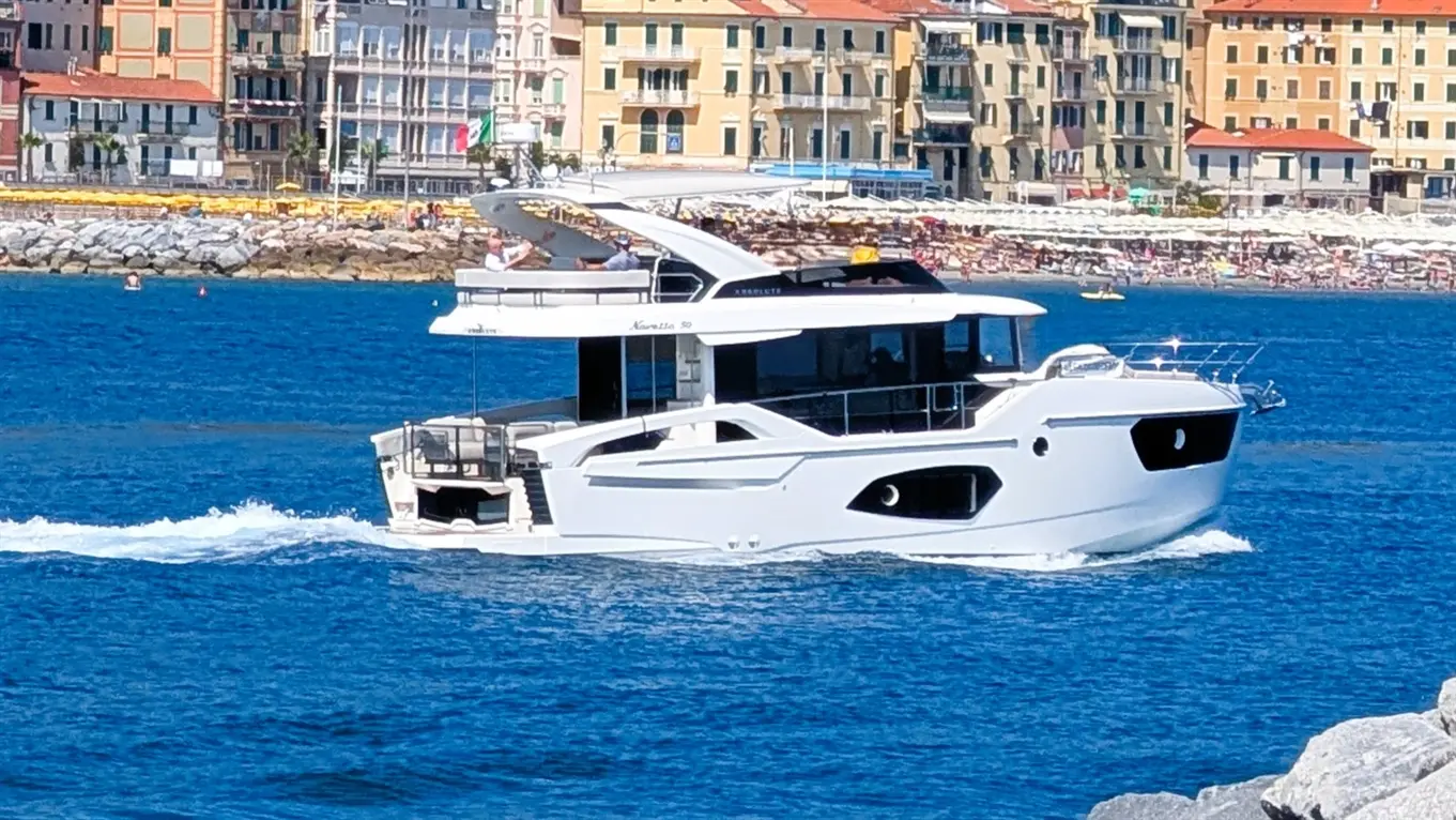 A sleek white motor yacht with a flybridge glides across blue water, with a beach and buildings visible in the background.