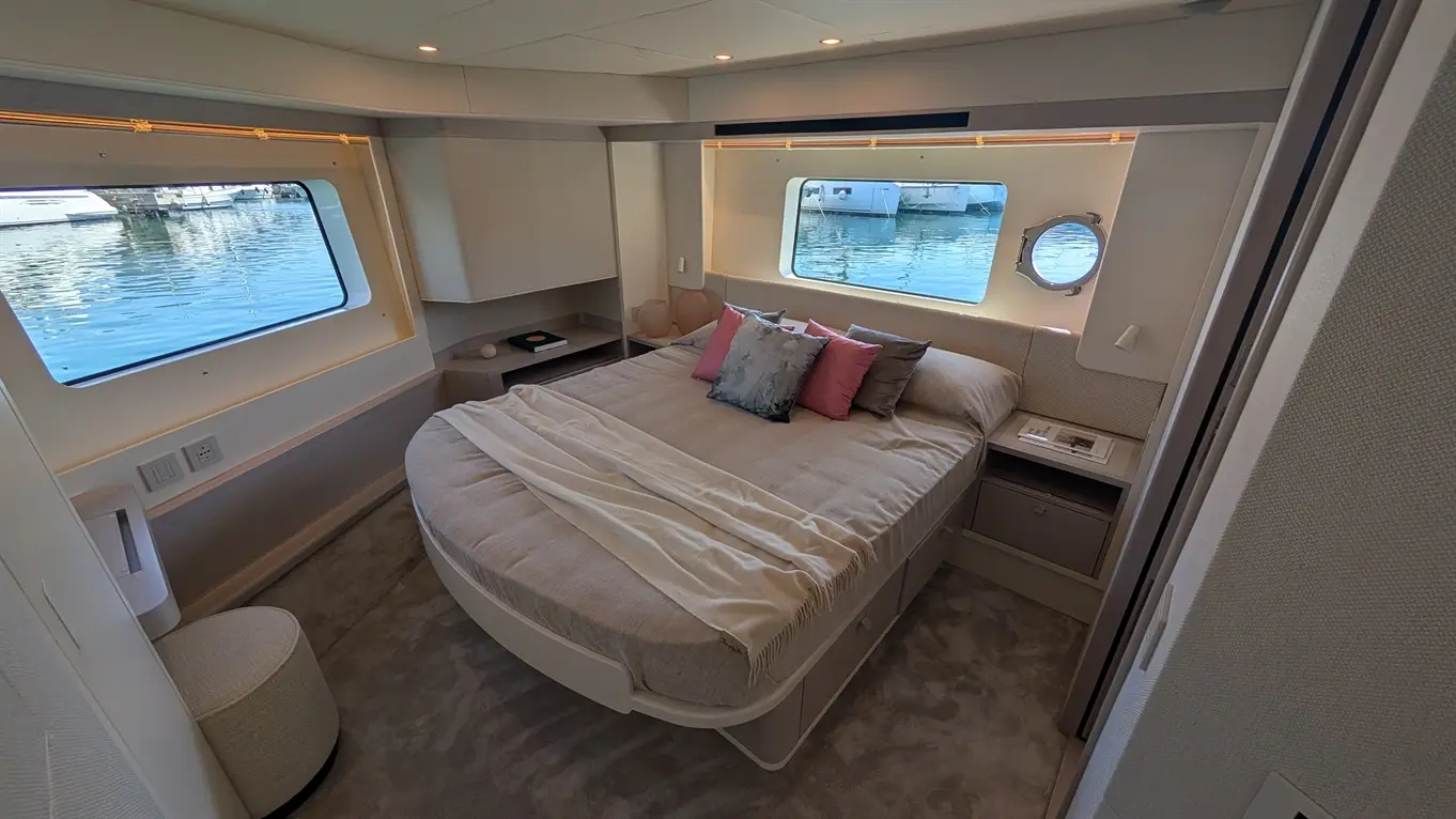 Luxury boat bedroom with circular bed, pink and grey pillows, and a window overlooking calm water and docked yachts.