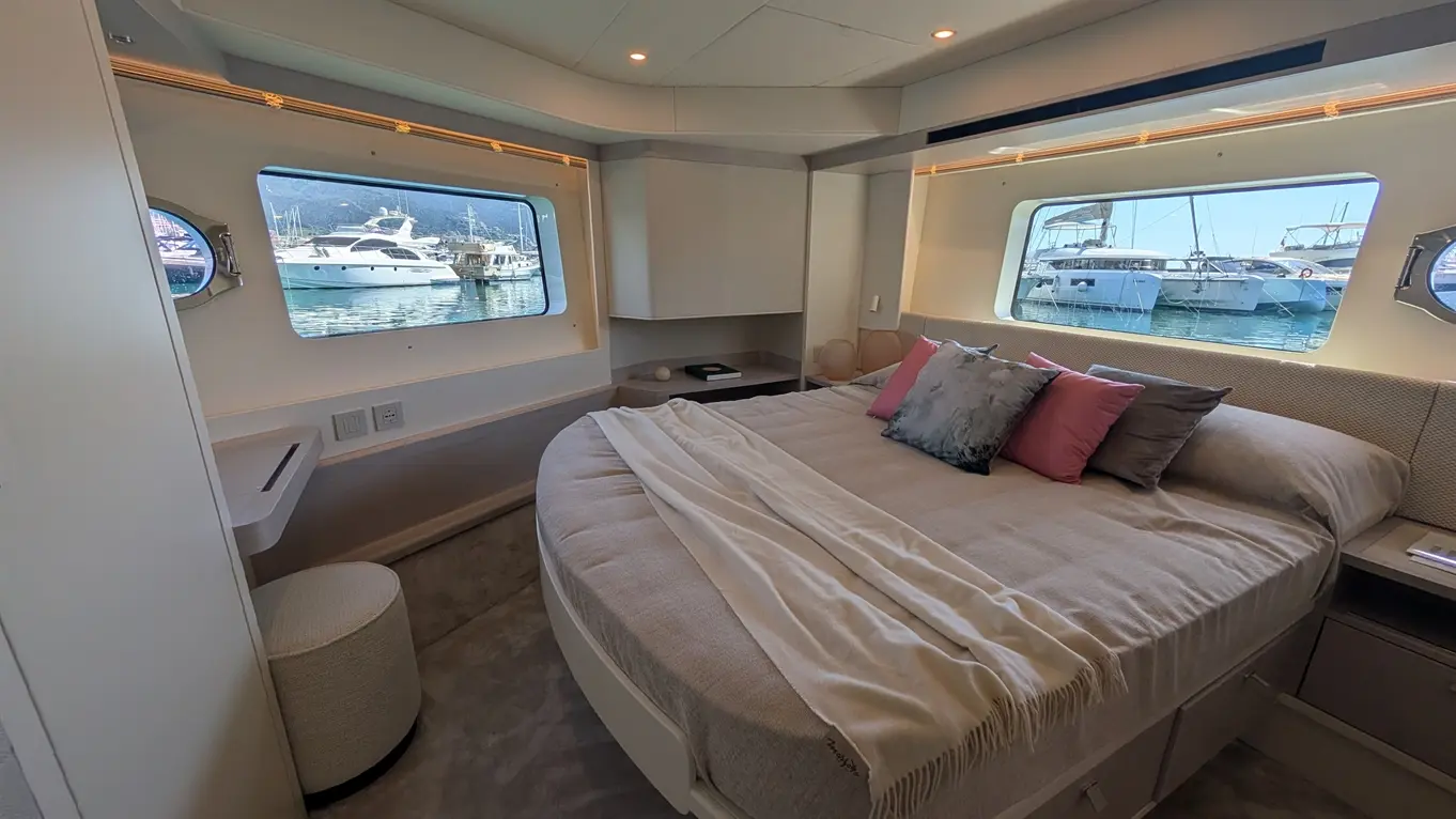 Luxury boat interior with round bed, decorative cushions, desk, and windows showcasing moored yachts outside.