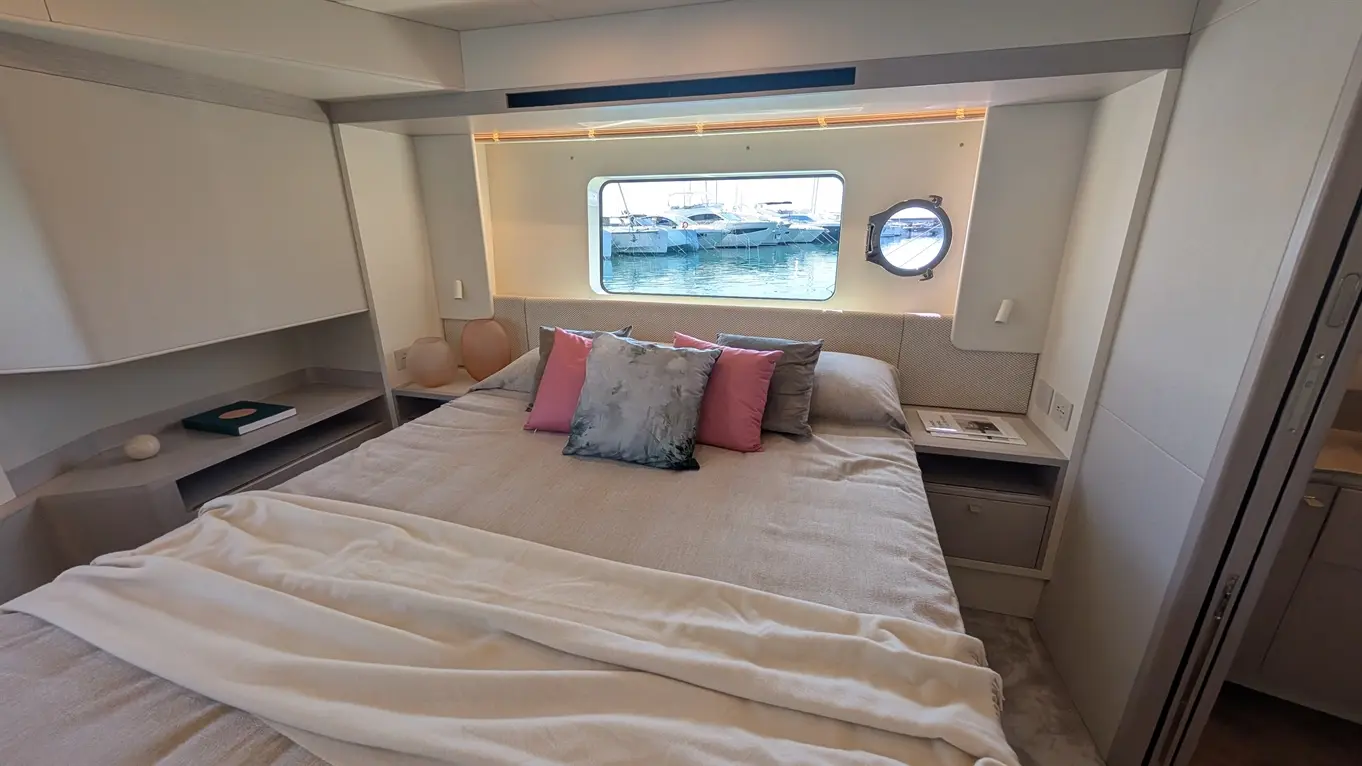 Stylish boat bedroom with a large bed, pink cushions, soft blanket, and port window showcasing docked boats outside.