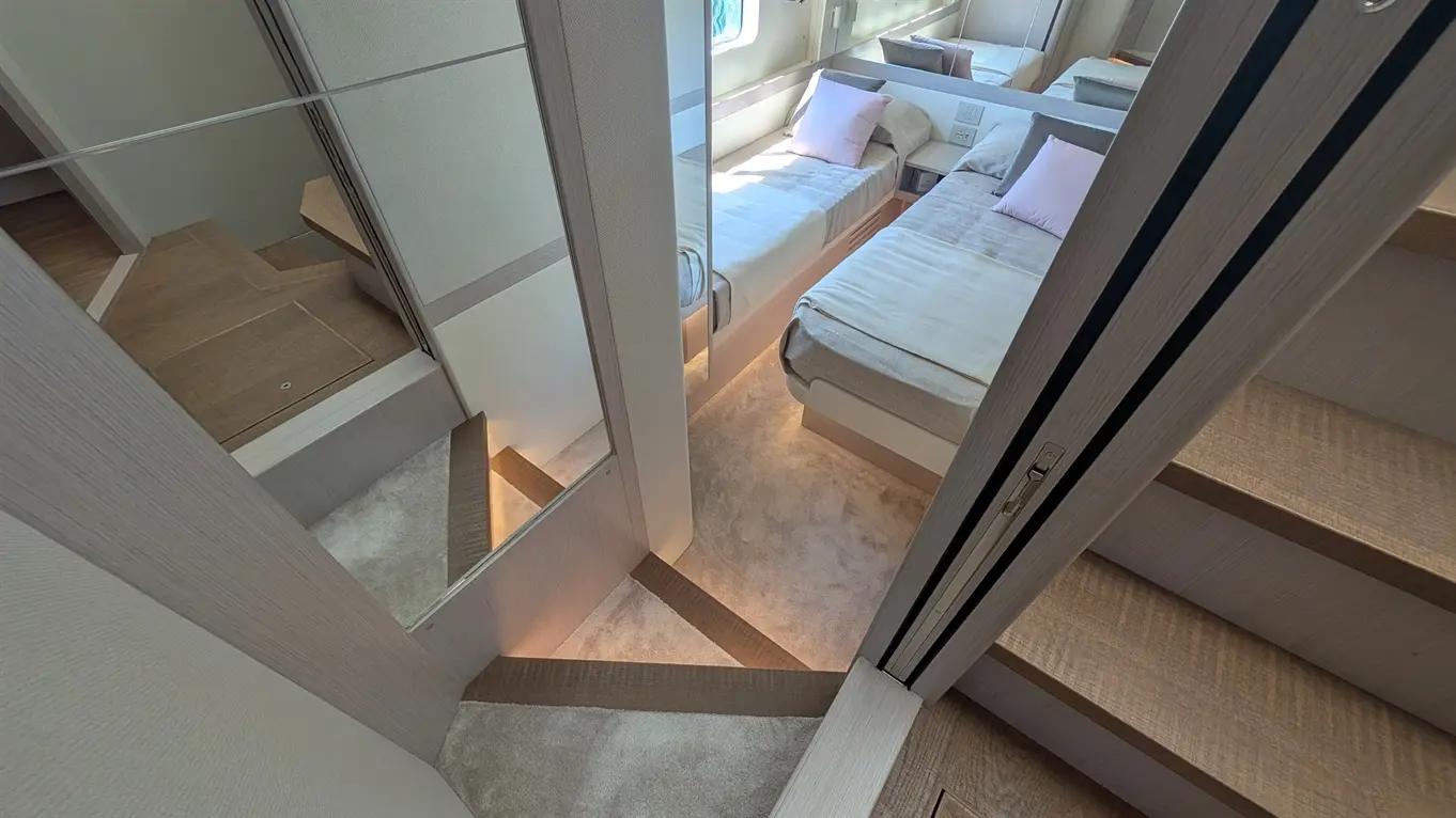 A modern, well-lit cabin area features two single beds, a mirror, and stylish wooden stairs leading to another level.