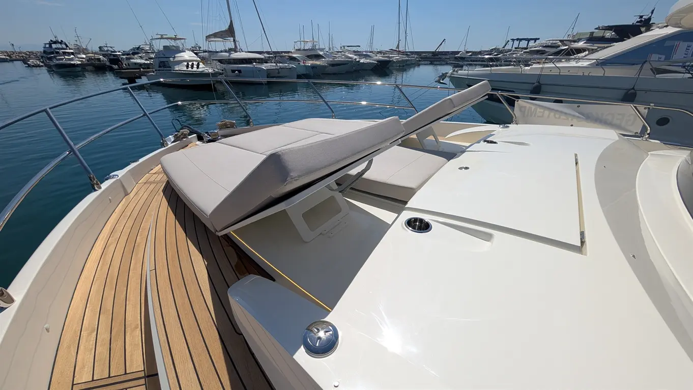 A luxury yacht's deck with a cushioned lounge, wooden flooring, and moored boats in the marina under clear skies.