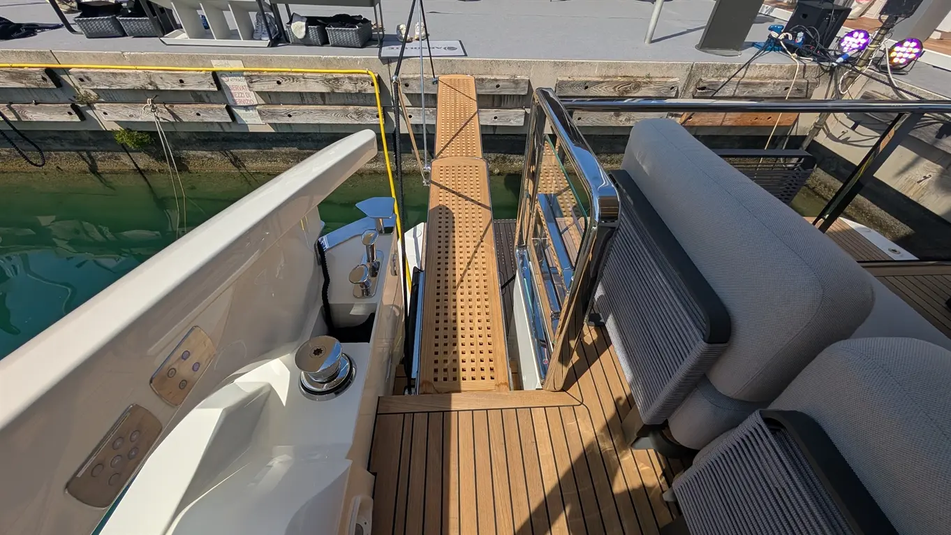 View of a boat's entrance ramp with wooden flooring leading to a dock, featuring a stainless-steel railing and seating.