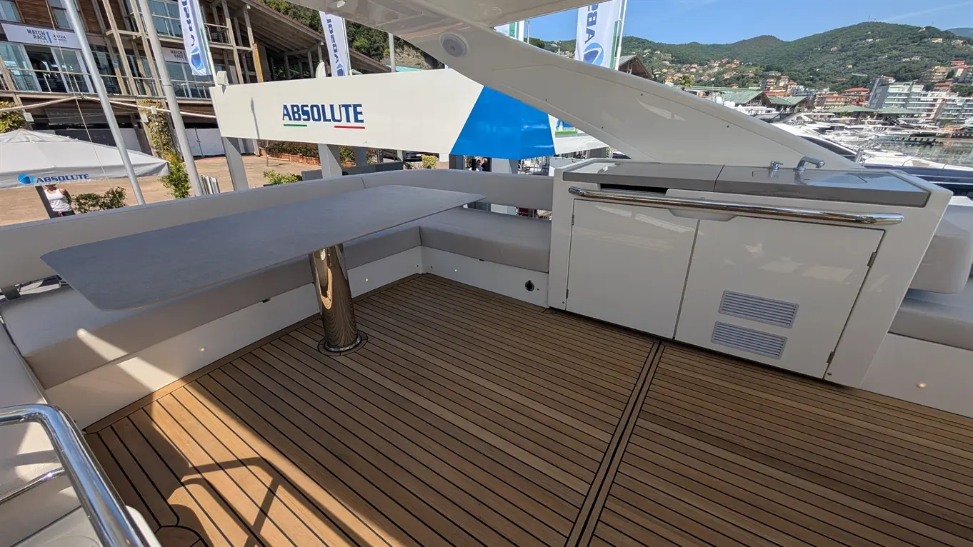 Spacious yacht deck featuring a grey table, plush seating, and a kitchenette among scenic marina views. "ABSOLUTE" branding visible.