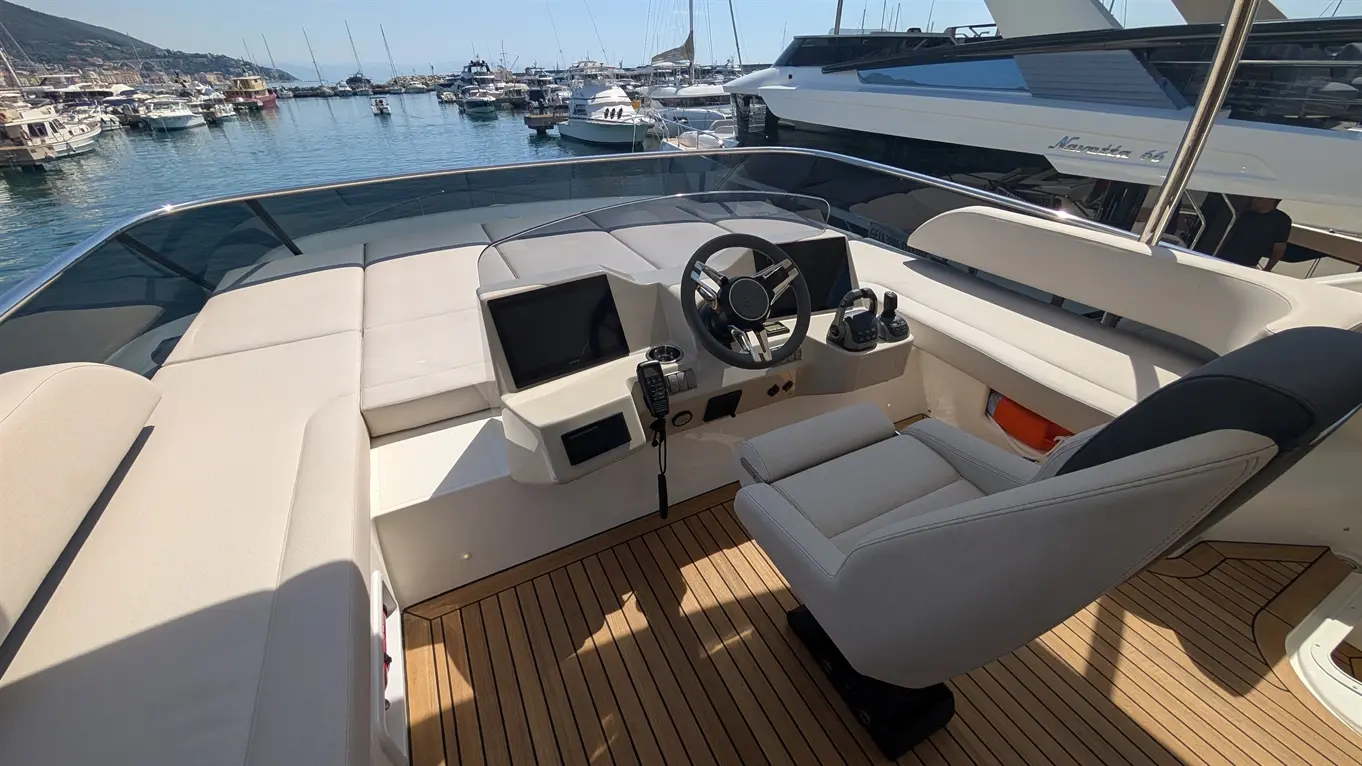Luxury boat interior featuring a steering wheel, dual screens, and plush seating on a wooden deck overlooking a marina.