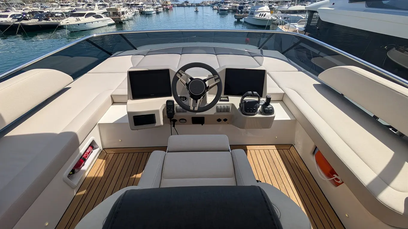 Luxury yacht cockpit with steering wheel, dual screens, and controls, overlooking a marina filled with boats on a sunny day.