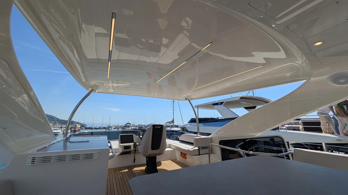 Modern yacht interior with panoramic views, showcasing a steering area, sleek furnishings, and docked boats in the background.