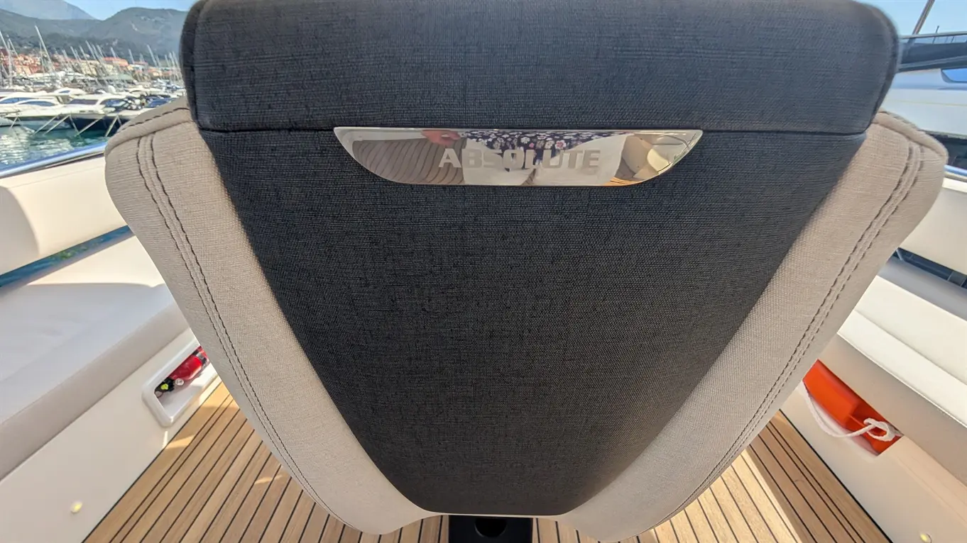 Back of a luxury boat seat with "ABSOLUTE" branding reflected, overlooking marina with yachts and scenic mountains.