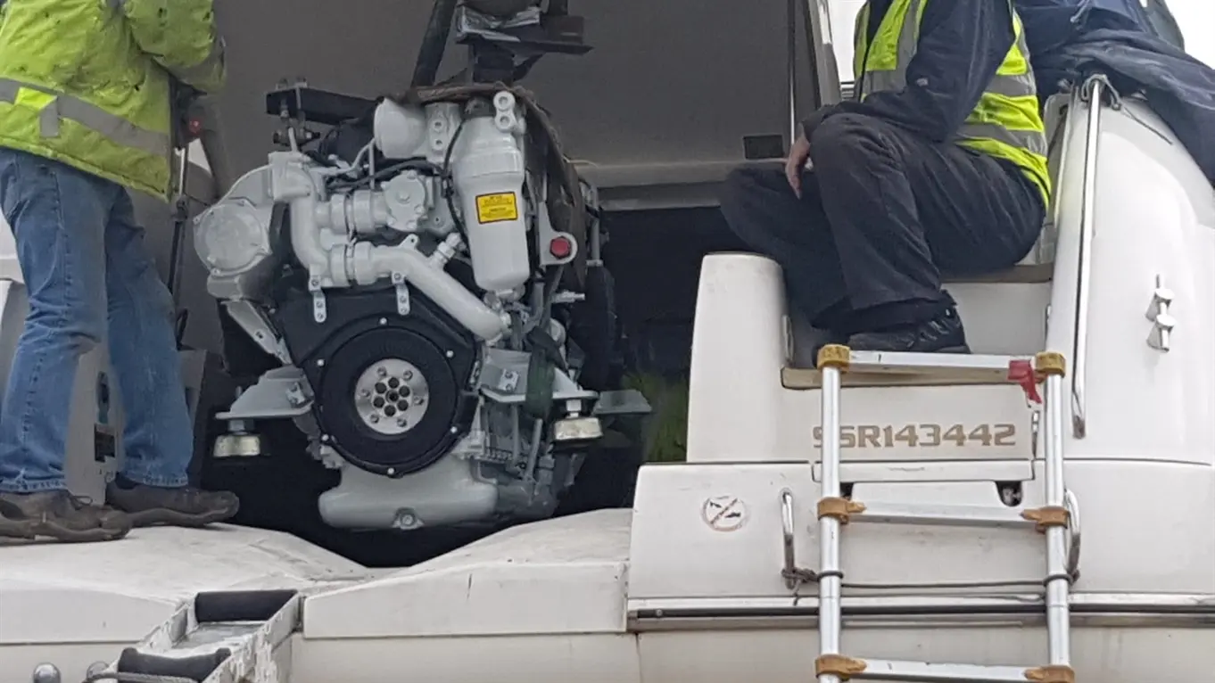 Two workers are installing an engine on a boat, with one worker standing and another seated on the steps, both wearing safety vests.