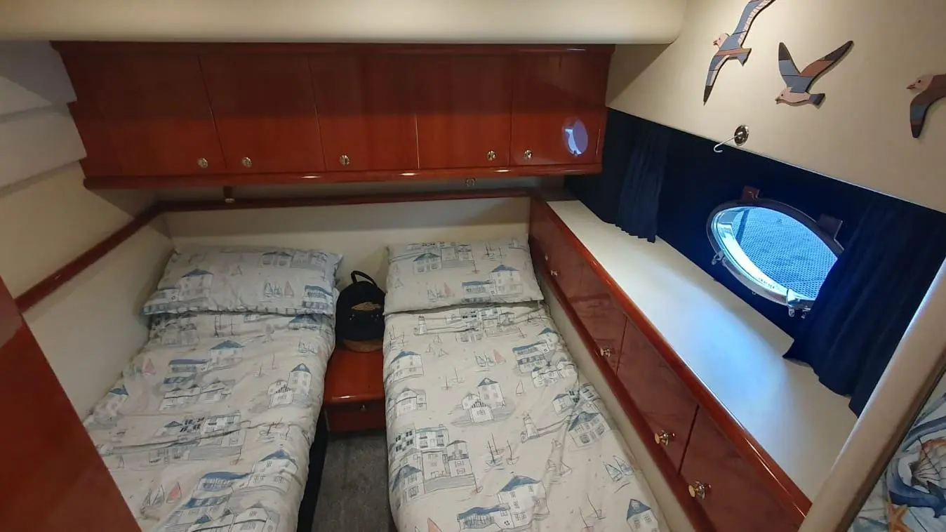 A compact cabin featuring two single beds with nautical-themed bedding, wooden storage above, and a porthole window.