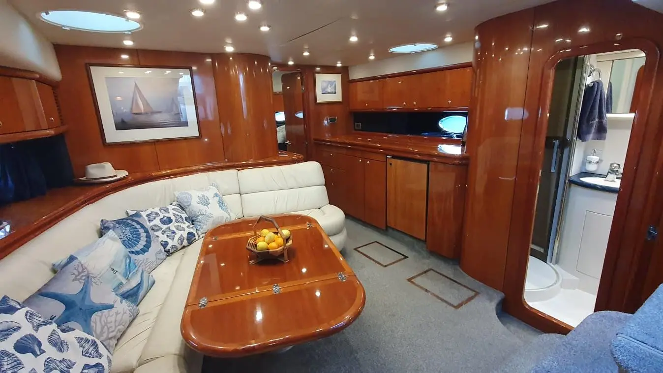 Luxury yacht interior featuring polished wooden surfaces, a cream leather sofa, decorative pillows, and a dining table with fruit.