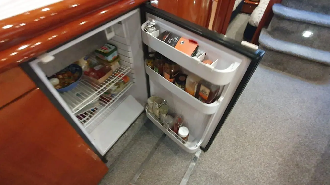 Open fridge in a boat kitchen, containing various food items, condiments, and vegetables on shelves. Carpeted floor visible.
