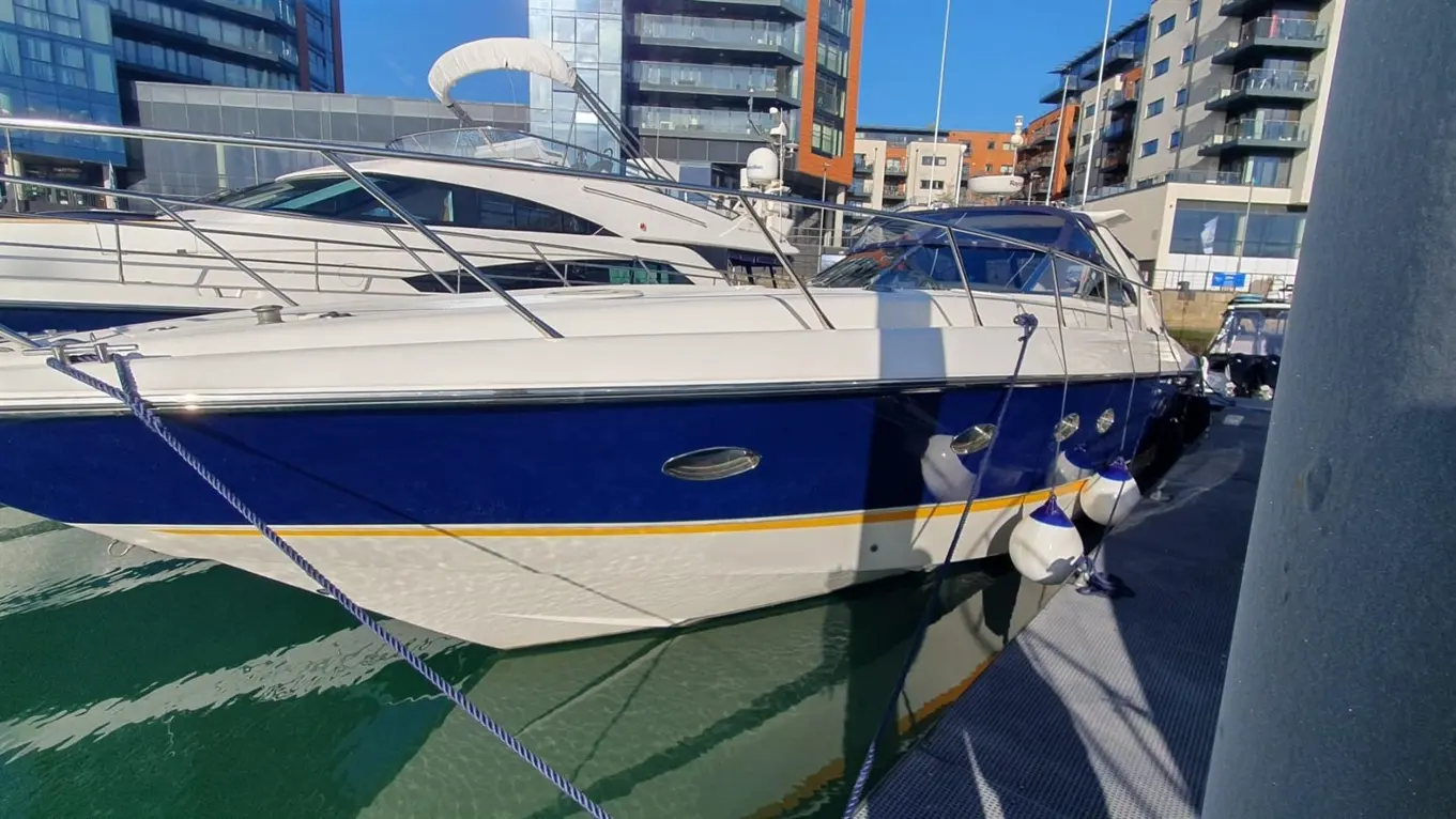 A blue and white motorboat is docked at a marina, surrounded by modern buildings and reflections on the water.