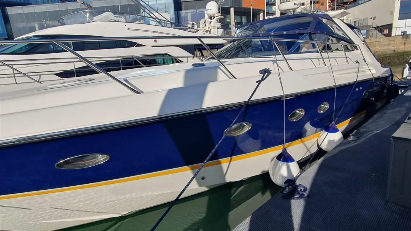 A luxury boat with a blue hull docked at a marina, featuring fenders, chrome details, and nearby modern buildings.