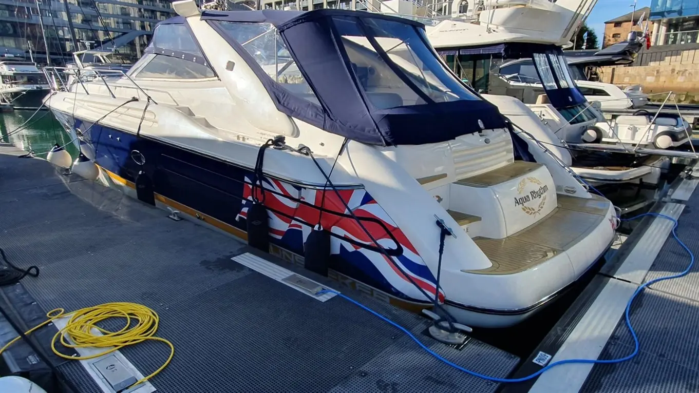 A moored boat featuring a Union Jack design, with wooden decking, alongside other vessels in a marina.