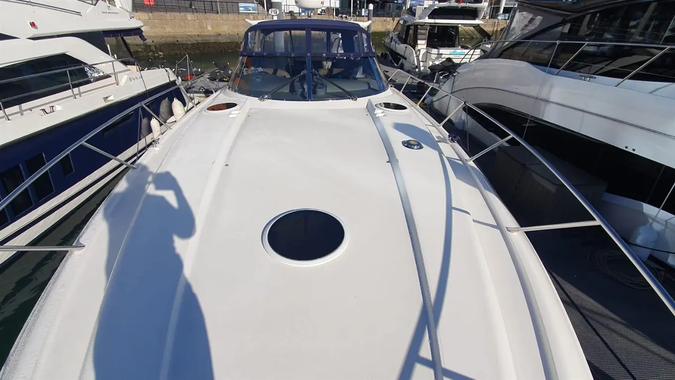 A white yacht's deck is shown, featuring a circular hatch and surrounding railings, with other boats docked nearby.