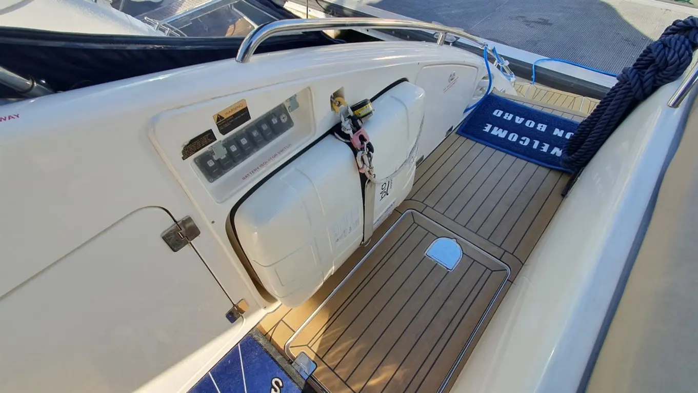 Close-up of a boat's control panel with a battery switch, storage compartment, and a welcome mat on the deck.