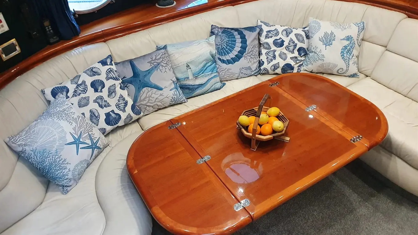 A cosy boat interior featuring a curved, cream leather sofa adorned with blue and white marine-themed cushions, and a fruit bowl.