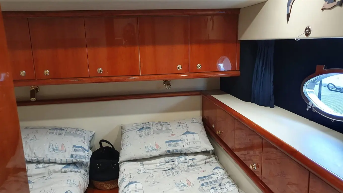 Interior of a boat cabin with two beds, wooden storage above, and a round porthole window, featuring nautical-themed bedding.