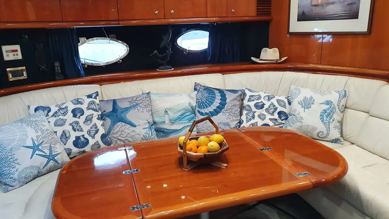 A boat interior featuring a circular wooden table, leather seating with blue-themed cushions, and a fruit basket.