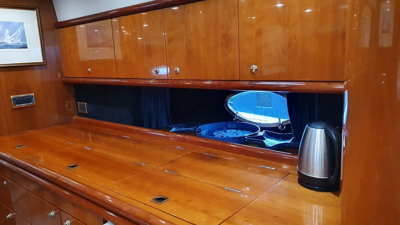 Interior of a luxury yacht featuring polished wooden surfaces, cabinets, a kettle, and a window with a view outside.