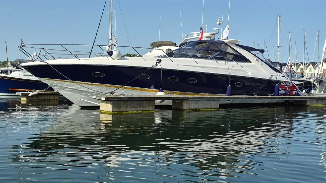 Luxury motor yacht docked at a marina, reflecting on calm water, with various boats and masts in the background.