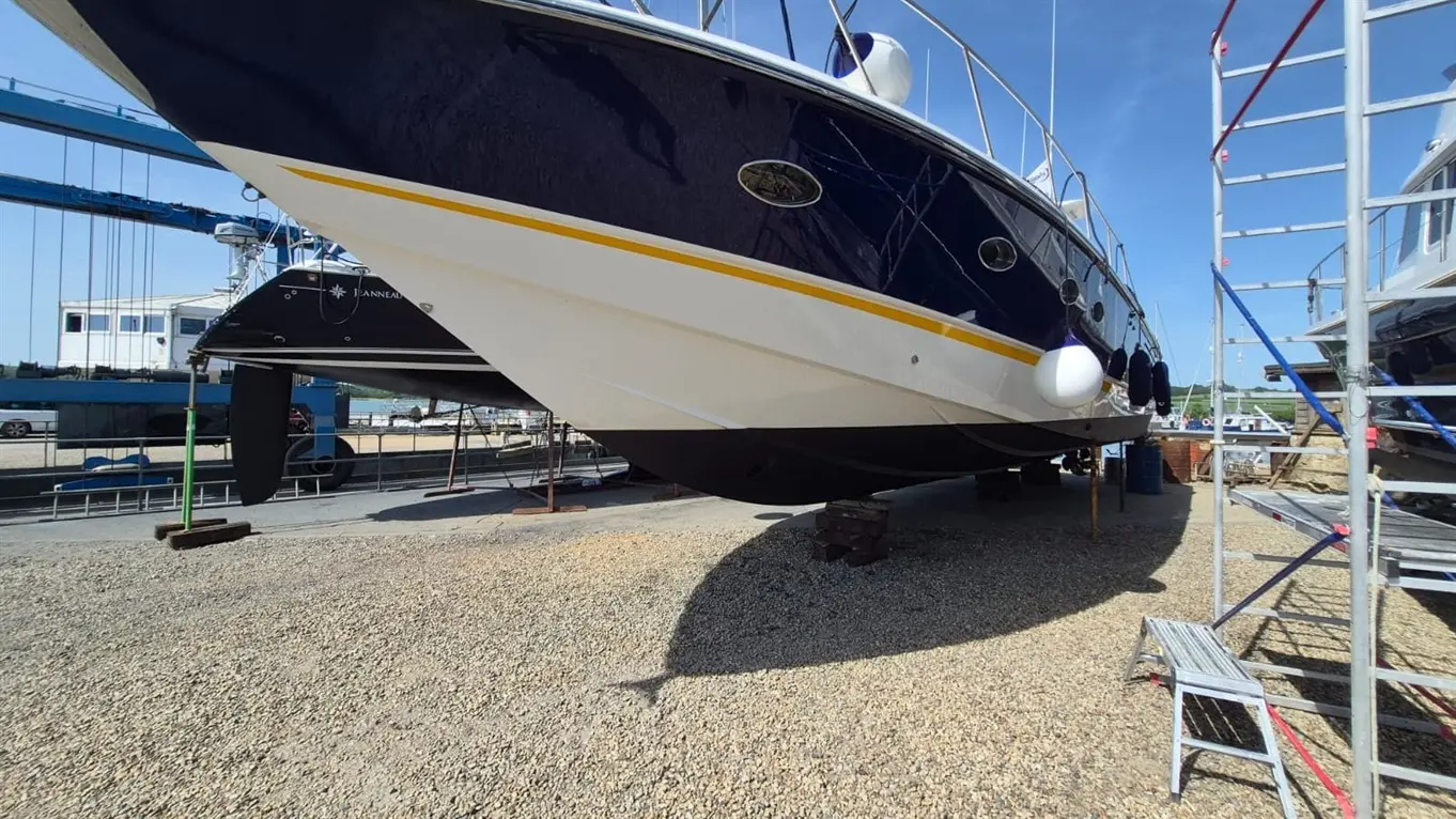 A blue and white yacht on a gravel surface, displayed on supports, with a portable ladder and workshop in the background.