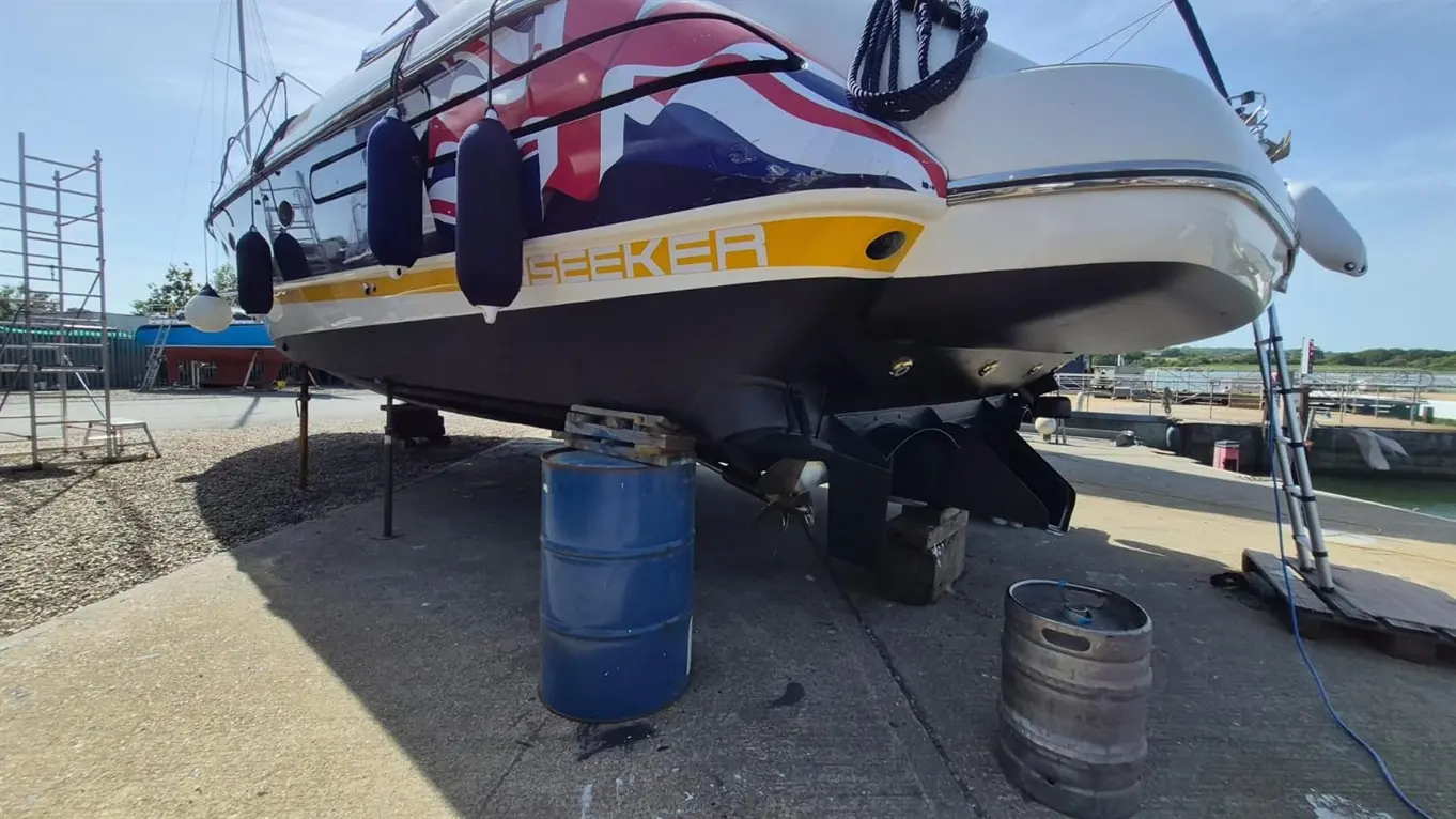 A boat named "Seeker" is on a dry dock, displaying British flag colours, with a scaffolding and barrels nearby.