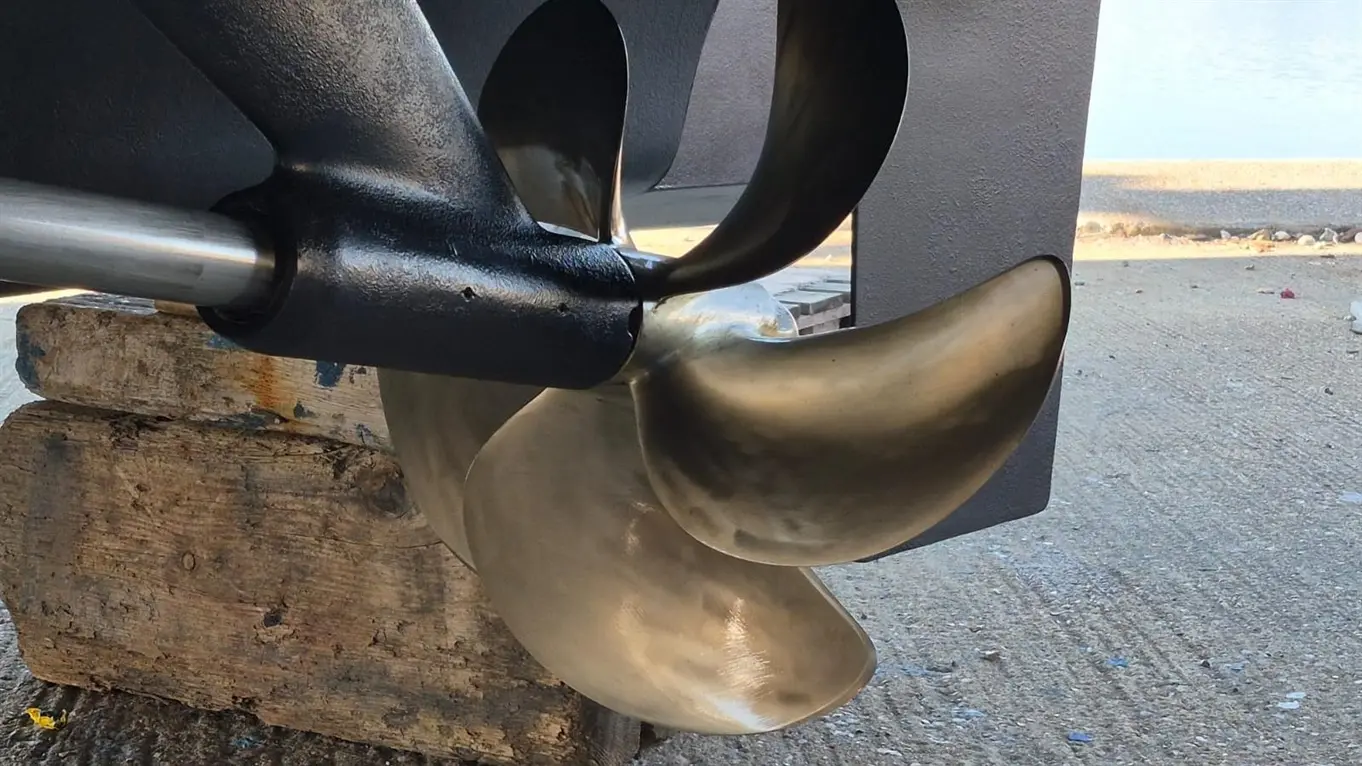 Close-up of a boat propeller attached to a shaft, resting on a wooden block with water and boats visible in the background.