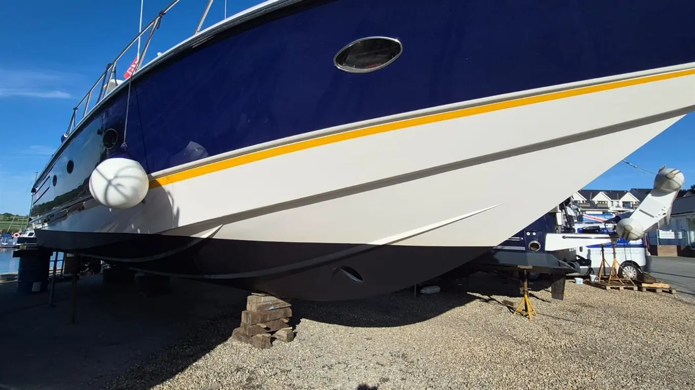 A large blue and white boat is lifted on supports, with visible fenders, the sky clear overhead, and a marina in the background.