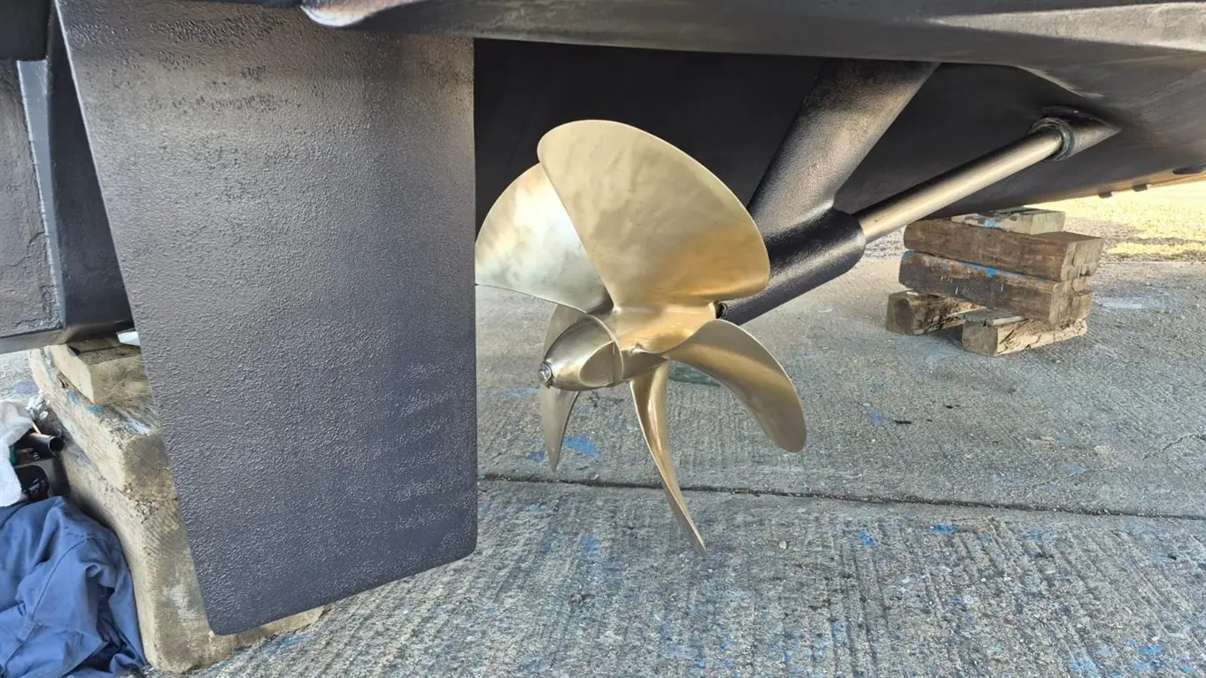 Bronze boat propeller and metal shaft visible beneath a vessel, supported on wooden blocks on a concrete surface.