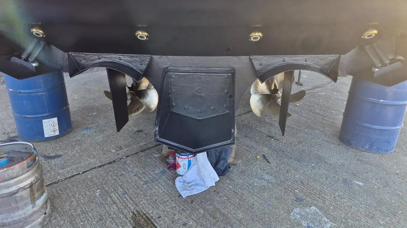 Boat propellers and steering gear mounted on a black hull, with two blue barrels and tools nearby on a concrete surface.