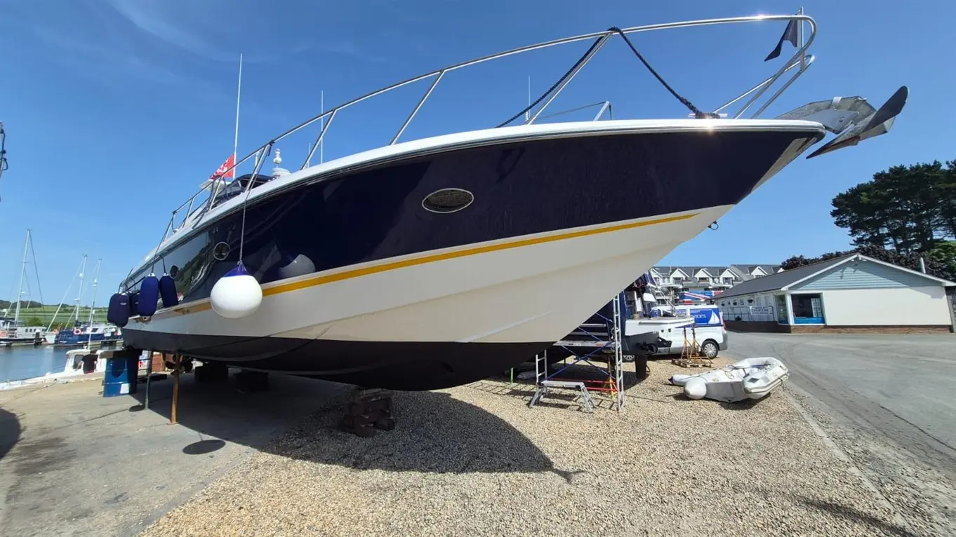 A large blue motorboat docked on a gravel surface in a marina, with visible boats and buildings in the background.