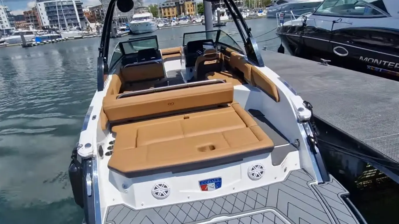 A luxury boat with tan seating and a sleek design, docked in a marina surrounded by other boats and buildings.