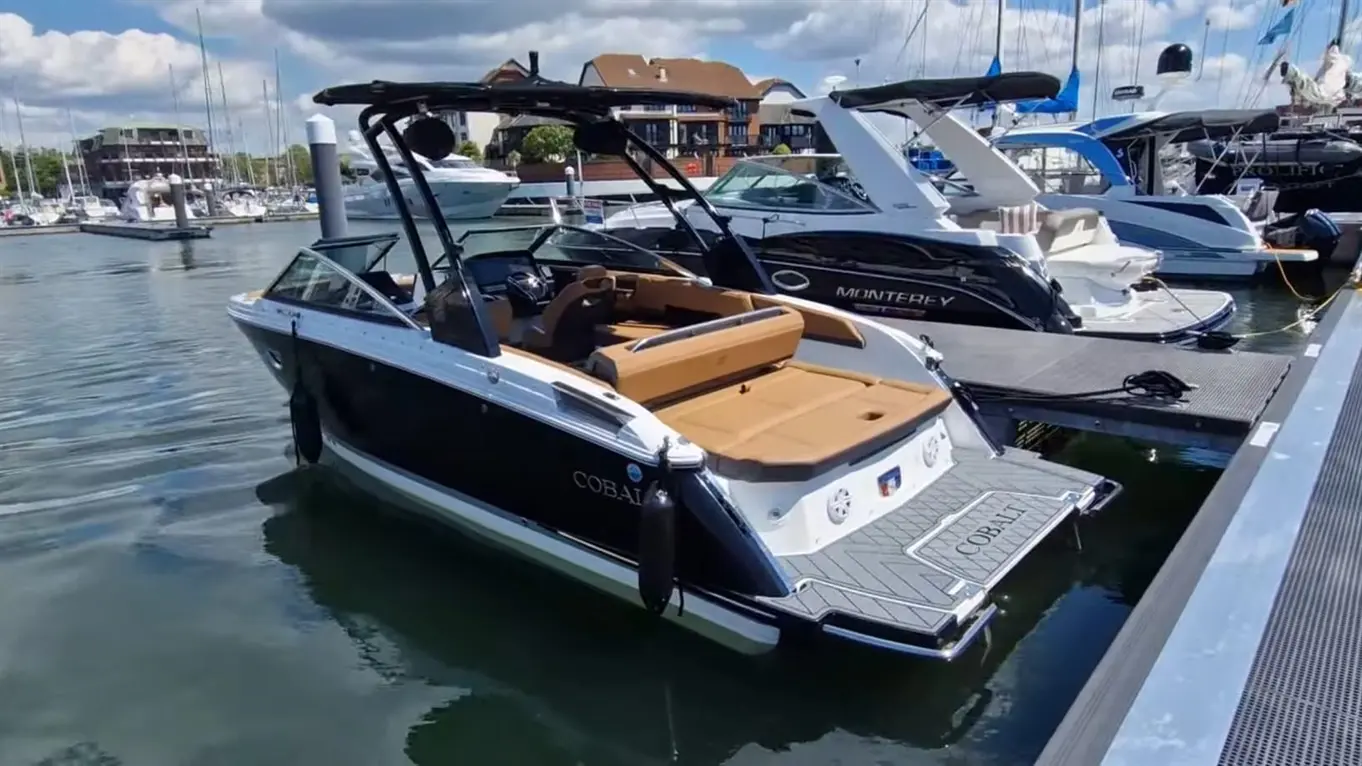 A Cobalt boat docked at a marina, featuring a brown interior, surrounded by multiple yachts and buildings under a partly sunny sky.