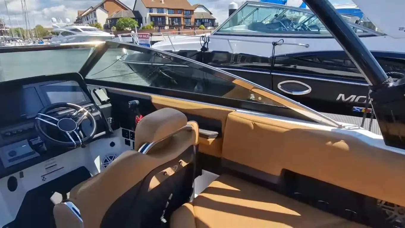 Interior view of a boat showing a steering wheel, control panel, and tan seating with a marina and other boats in the background.