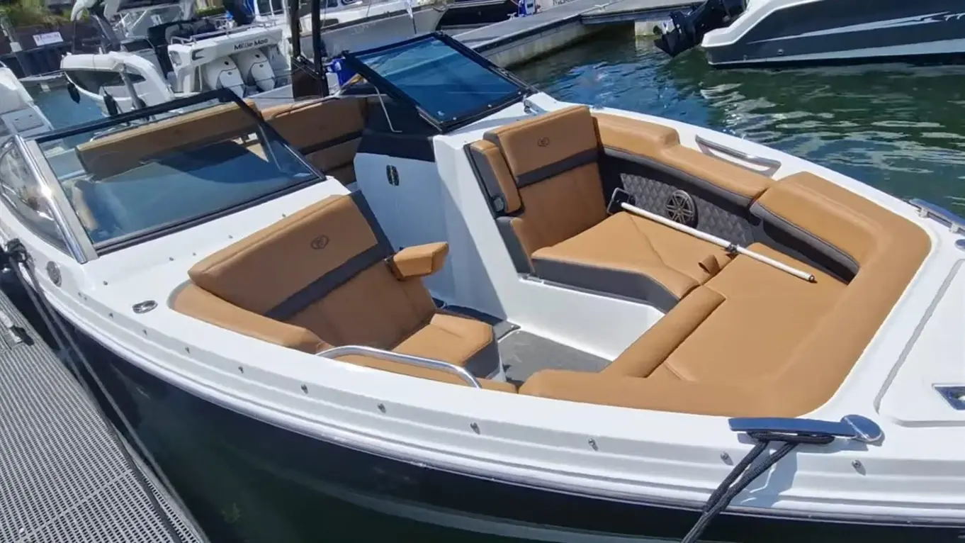 Modern boat with brown leather seating and a glass windshield, docked by a marina with other boats in the background.