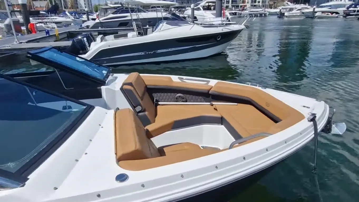A modern boat with beige seating is docked in a marina, surrounded by other boats and reflecting water.