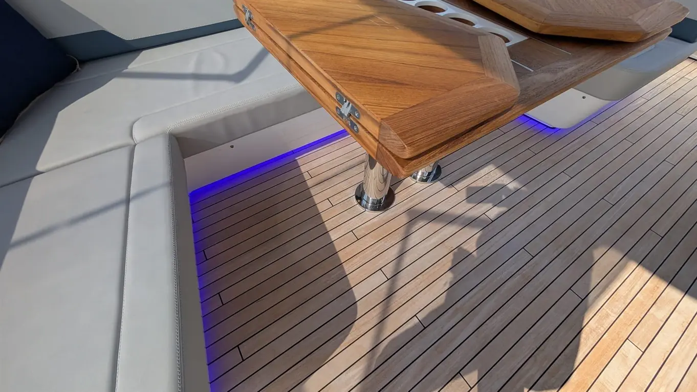 Wooden table on a boat deck with stainless steel supports, surrounded by wooden flooring and illuminated by blue LED lights.