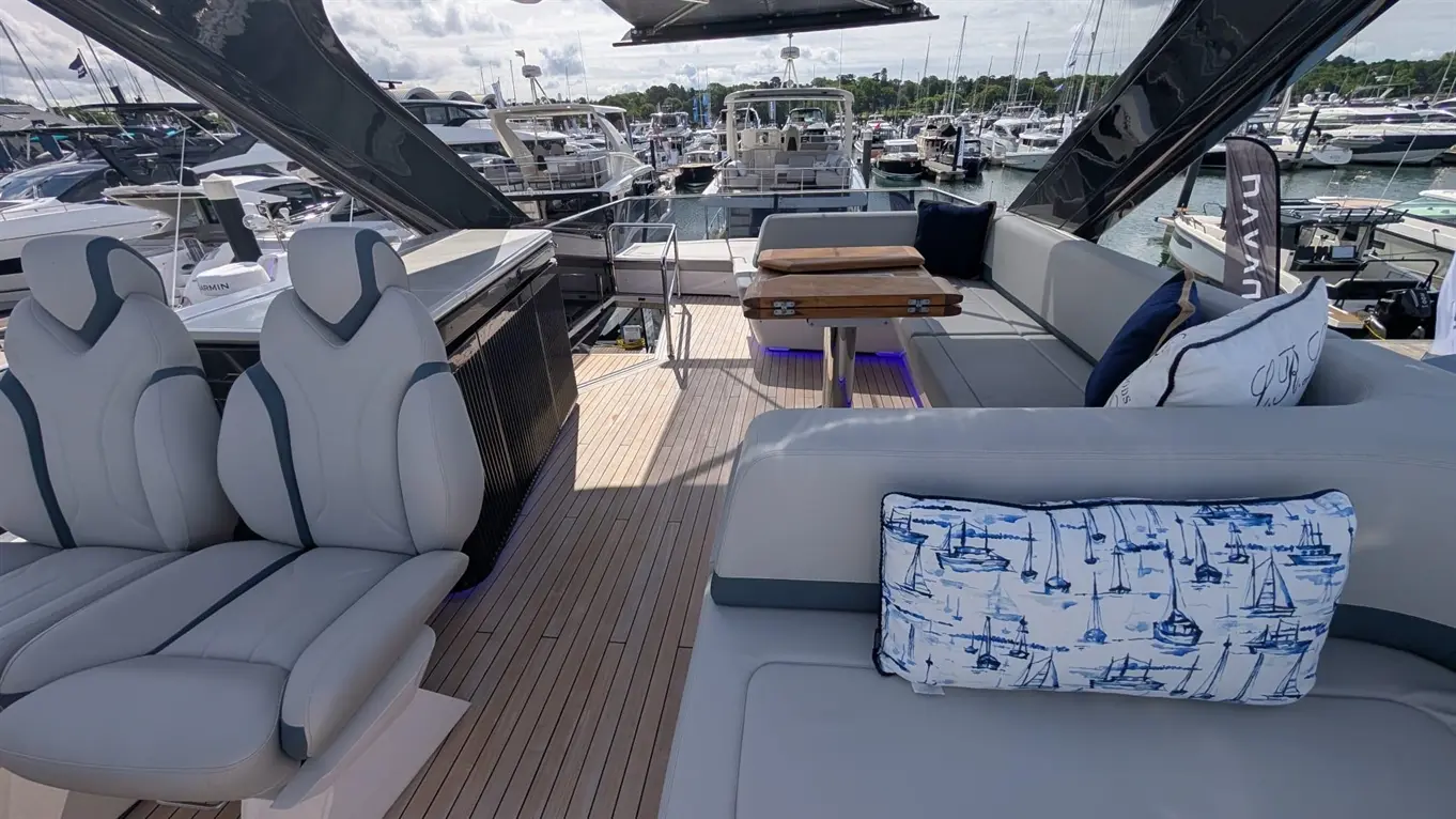 Stylish boat interior with grey seating, wooden table, and nautical-themed cushions, overlooking a marina filled with yachts.