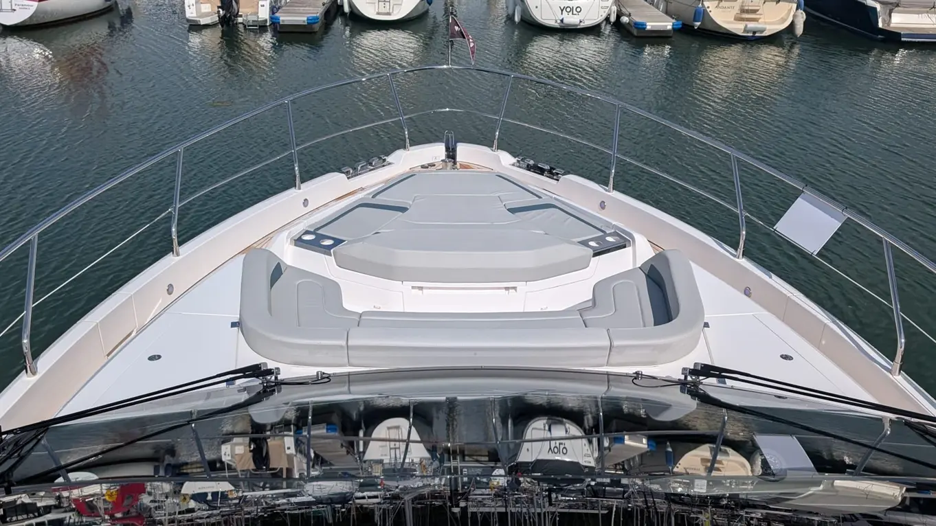 A yacht's bow features a grey seating area, overlooking a marina with moored boats and a calm water surface.