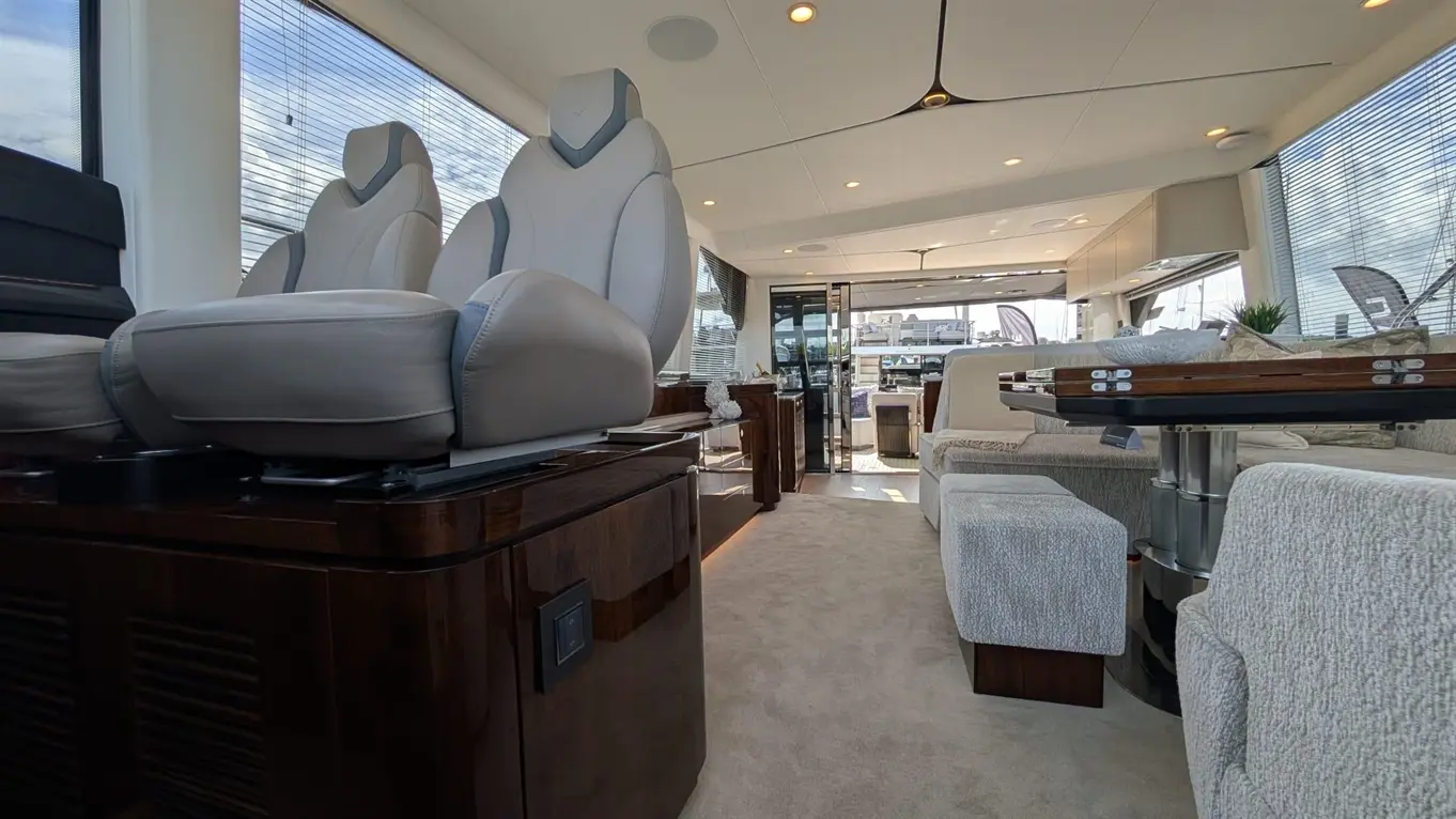Interior of a luxurious yacht with plush grey seats, wooden furnishings, and a dining area, featuring large windows and soft lighting.