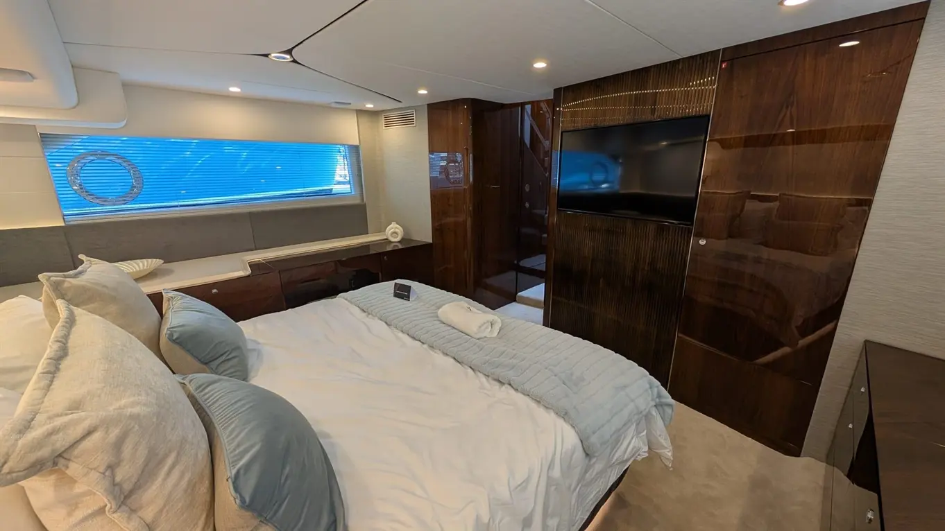 Luxury yacht bedroom featuring plush bedding, a TV, and ocean views through partially open blinds. Elegant decor in warm tones.