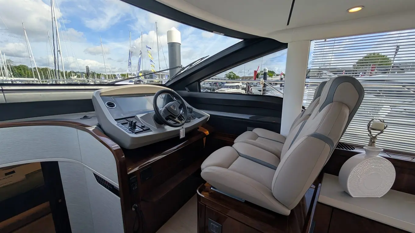 Interior of a luxury yacht showing a steering wheel, control panel, two beige seats, and decorative vase with sailing boats outside.