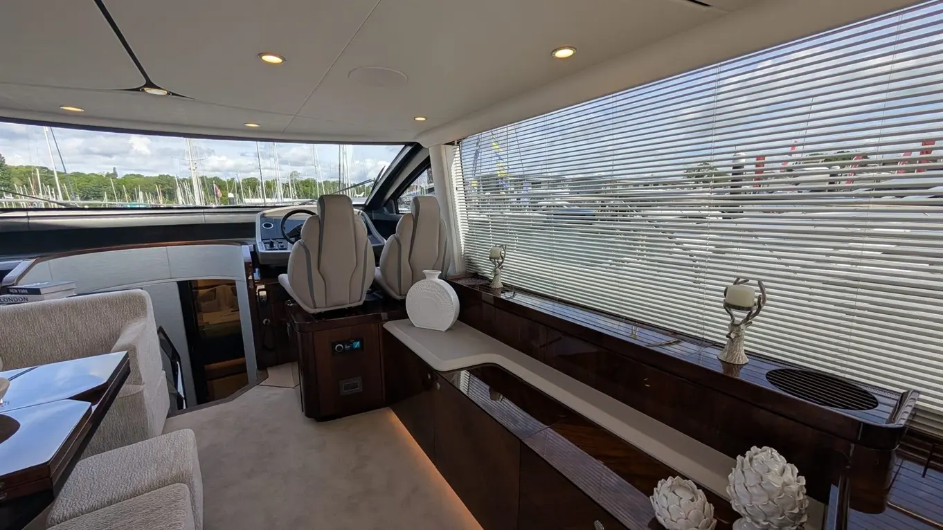 Interior of a luxury yacht featuring grey seating, a steering console, decorative vases, and views of moored boats through blinds.