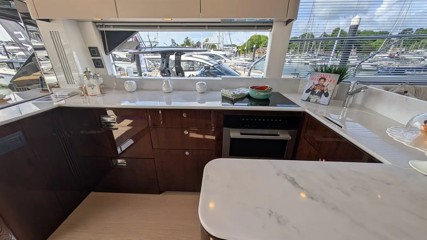 Modern kitchen onboard a yacht, featuring a marble countertop, oven, and a view of the marina, with a cooking magazine visible.